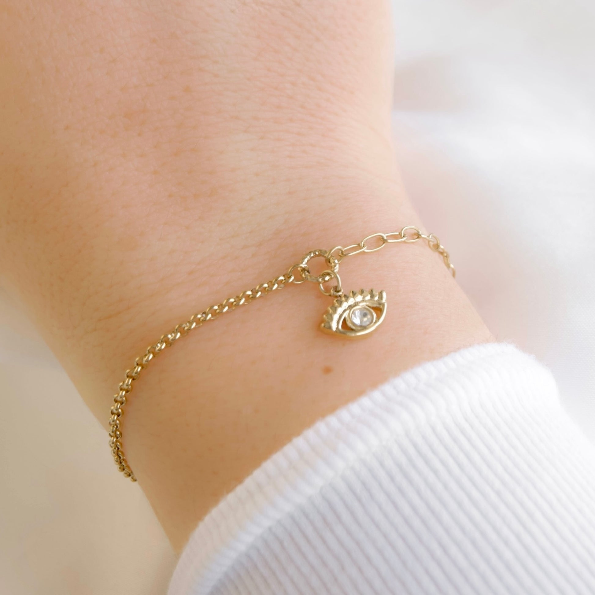 A delicate gold chain bracelet with an evil eye charm, shown worn on a person's wrist over a white ribbed long-sleeve top.