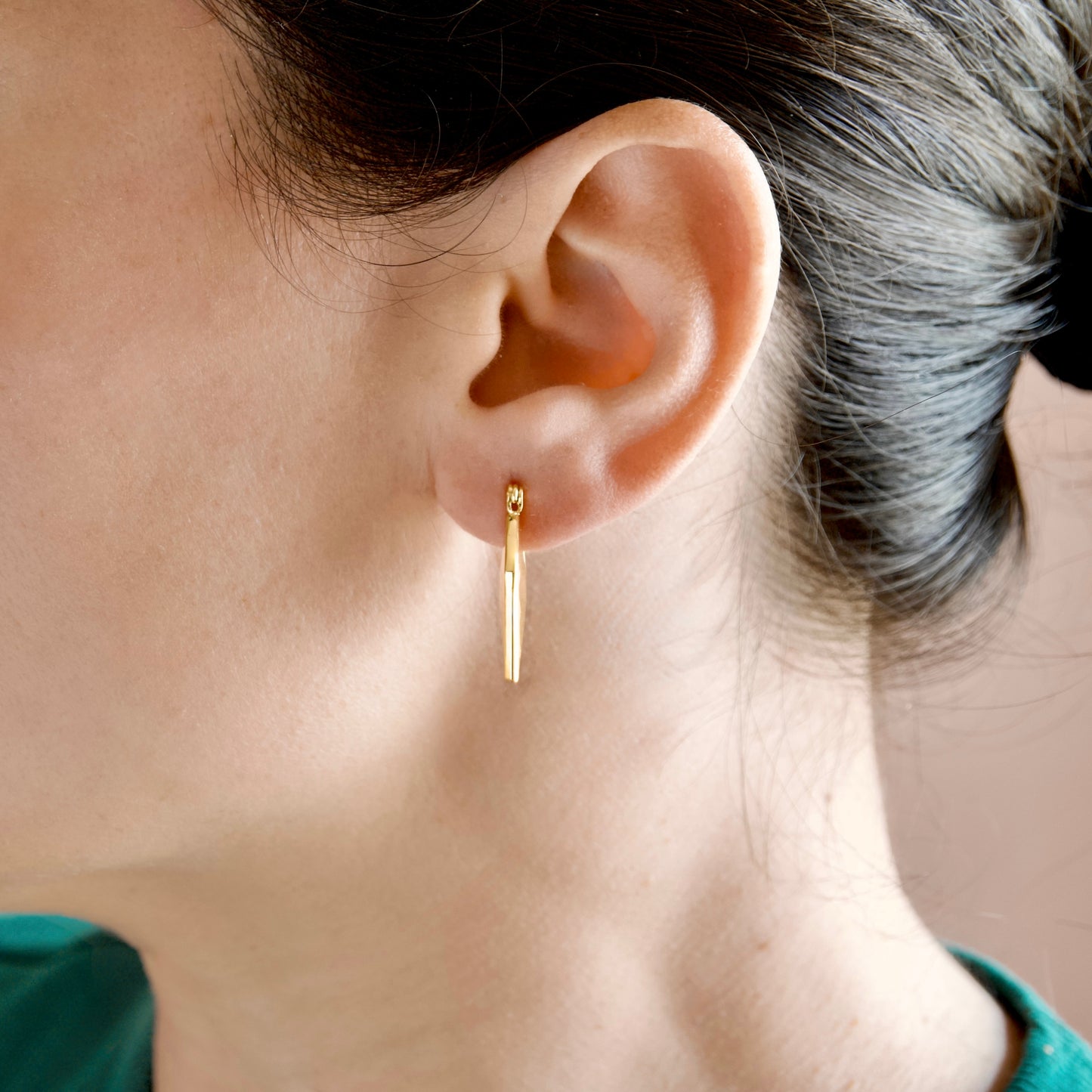 A close-up of a person's ear wearing a delicate gold drop hoop earring with a long, thin drop element.