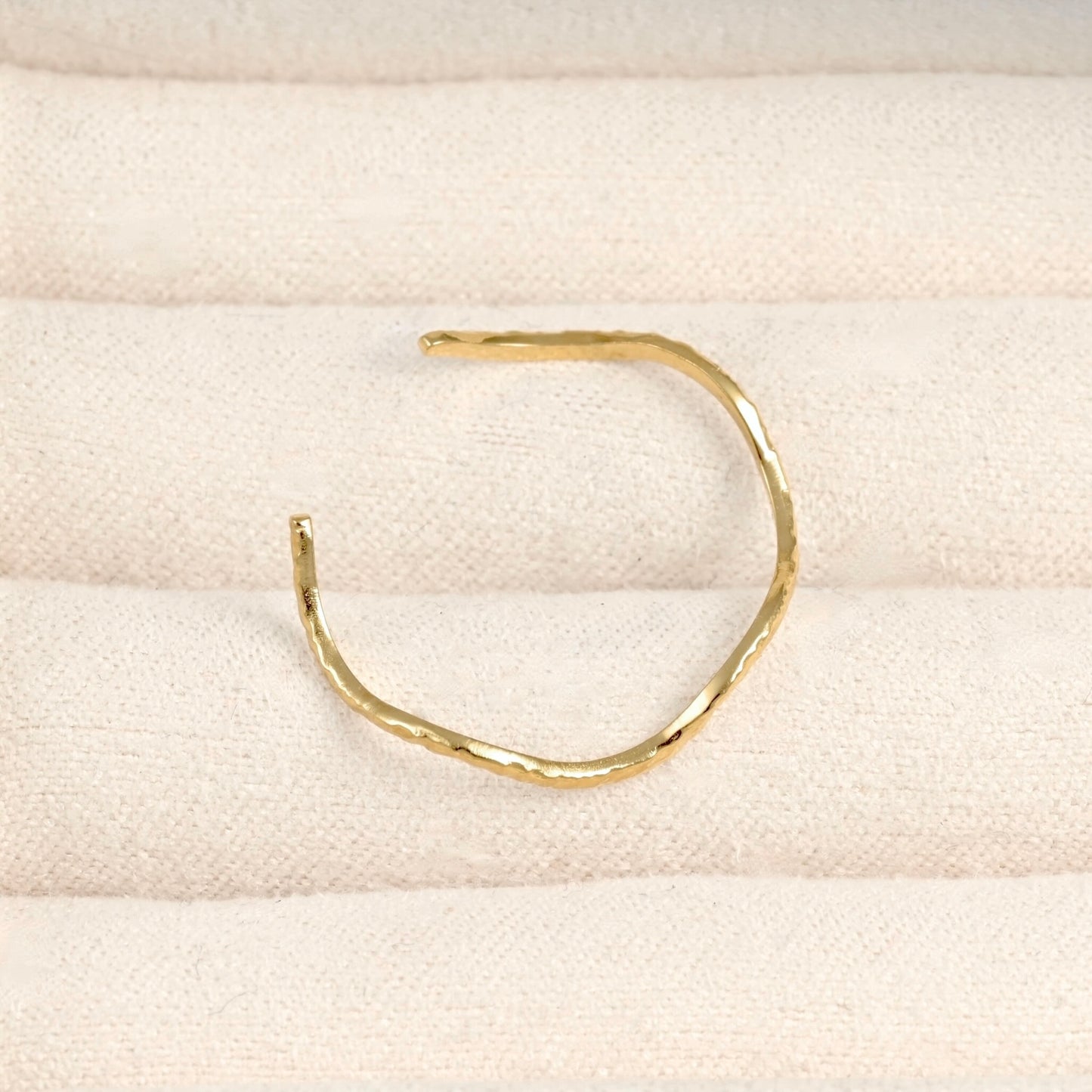 A single hammered gold open cuff ring displayed on a textured cream fabric surface.