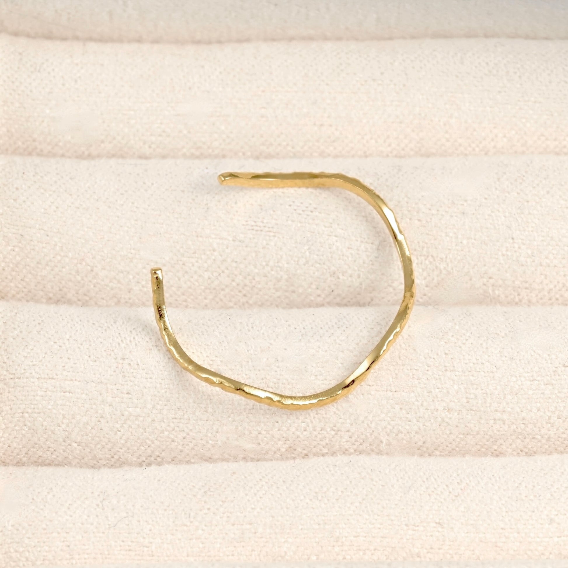 A single hammered gold open cuff ring displayed on a textured cream fabric surface.