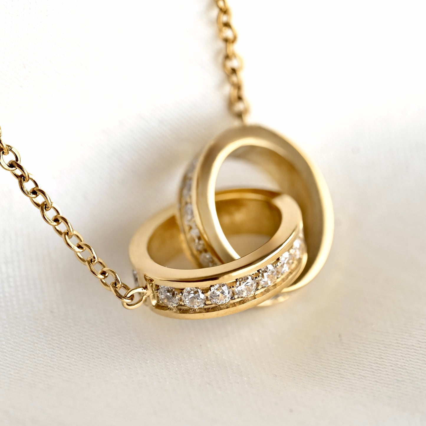 A gold pendant necklace featuring two interlocking rings adorned with a row of clear pavé diamonds, resting on a white textured fabric background.