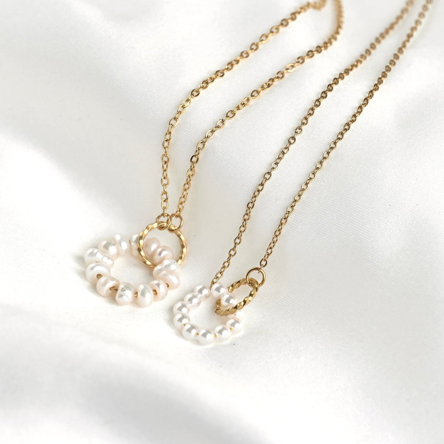 Two delicate gold chain necklaces with small pearl-bead circular pendants, arranged on a soft white satin fabric background.