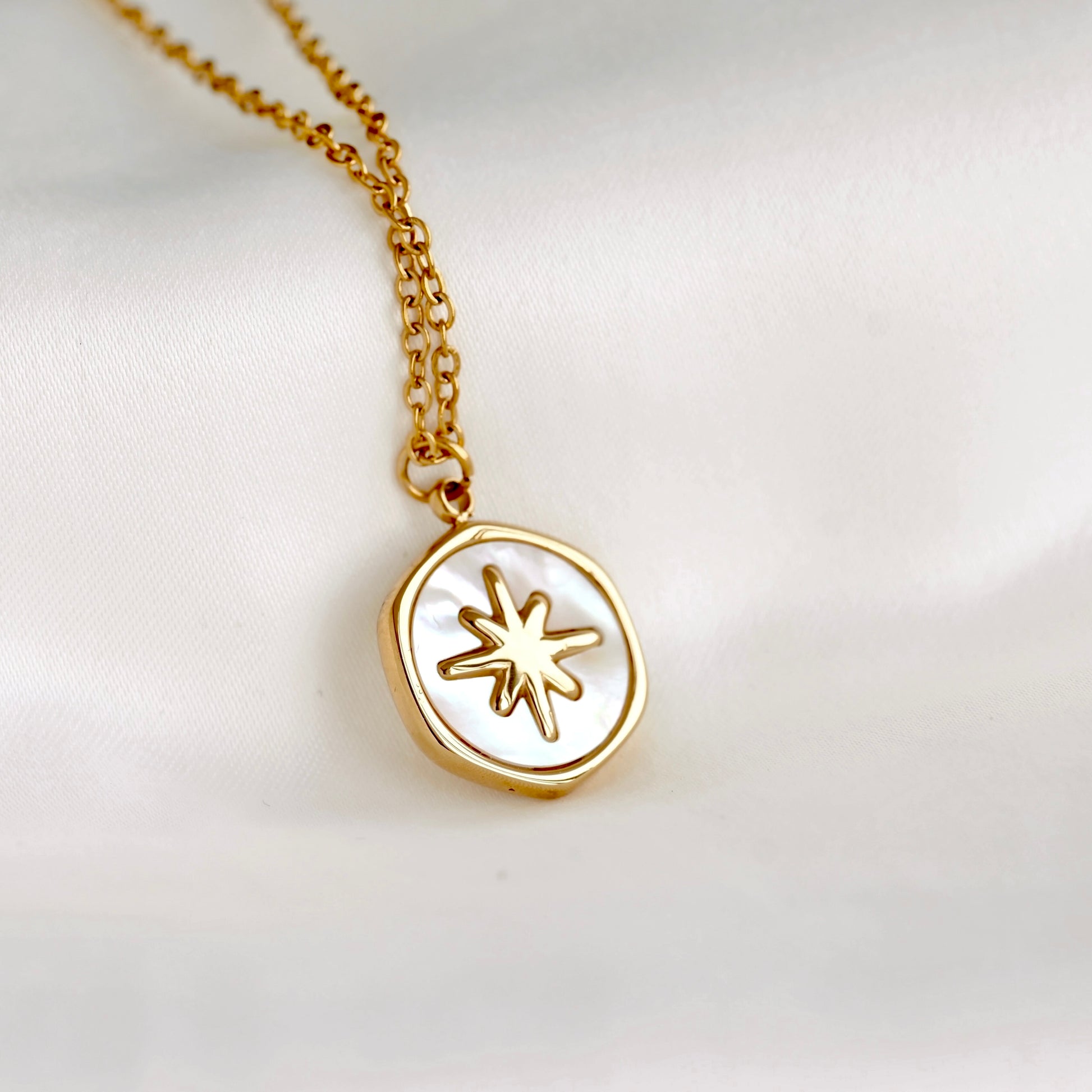 A gold chain necklace with a round pendant featuring a white mother-of-pearl background and a gold starburst design, displayed on a soft white fabric background.