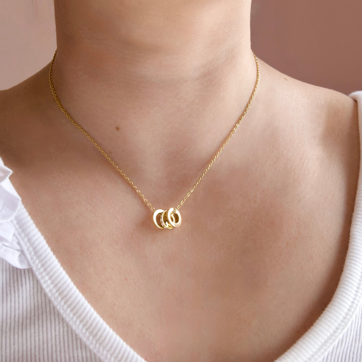 A close-up of a person wearing a delicate gold chain necklace with a small interlocking rings pendant, paired with a white ribbed top against a soft pink background.