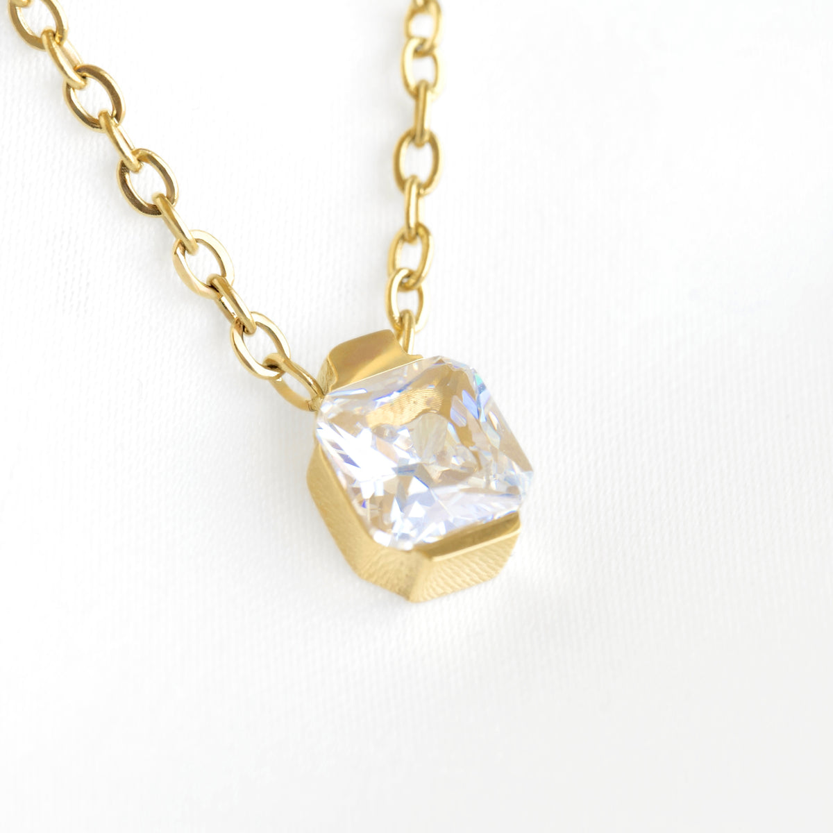 A gold chain necklace with a faceted geometric gold pendant holding a clear, cushion-cut gemstone, shown on a clean white background.