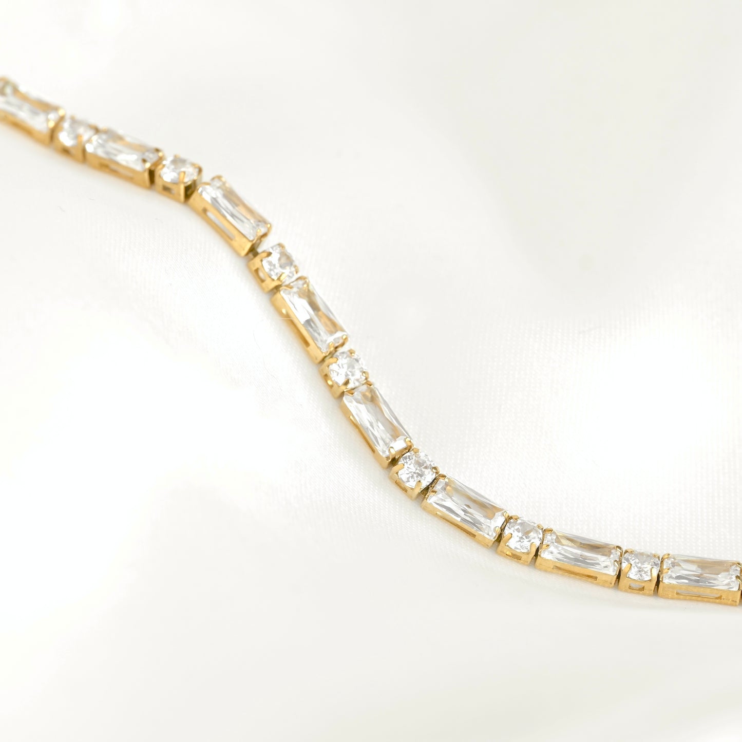 A gold tennis bracelet featuring mixed baguette and round cut clear stones, photographed on a soft white satin fabric background.