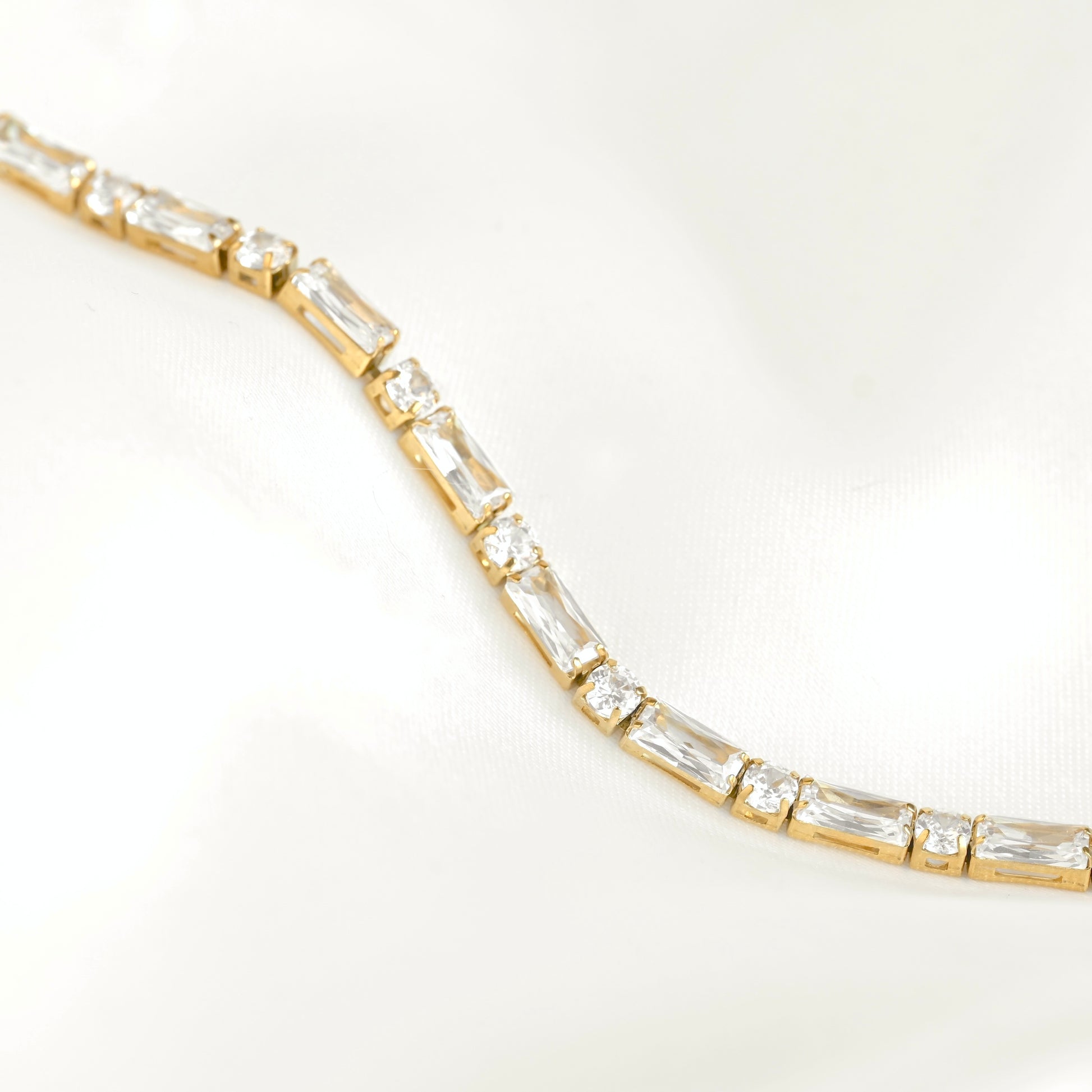 A gold tennis bracelet featuring mixed baguette and round cut clear stones, photographed on a soft white satin fabric background.