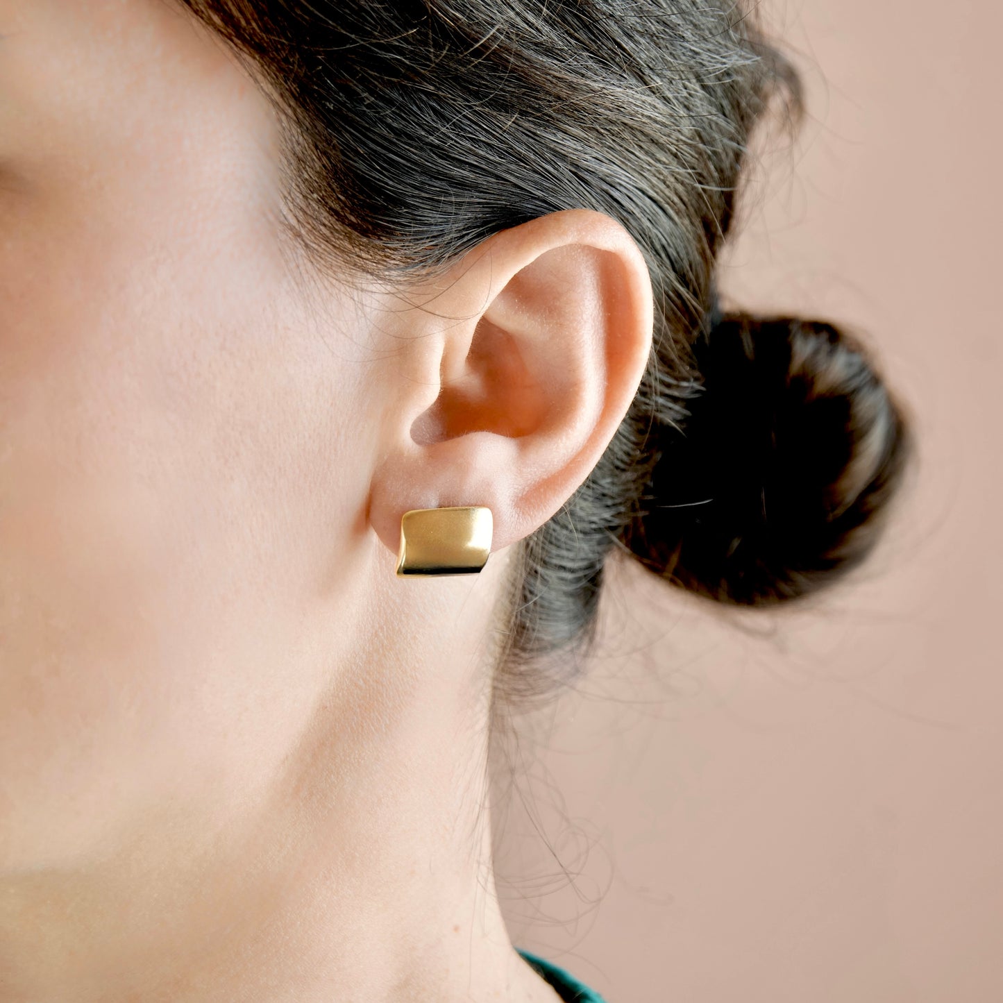 A close-up of a woman's ear wearing a small, square gold stud earring with a polished finish, set against a soft beige background.