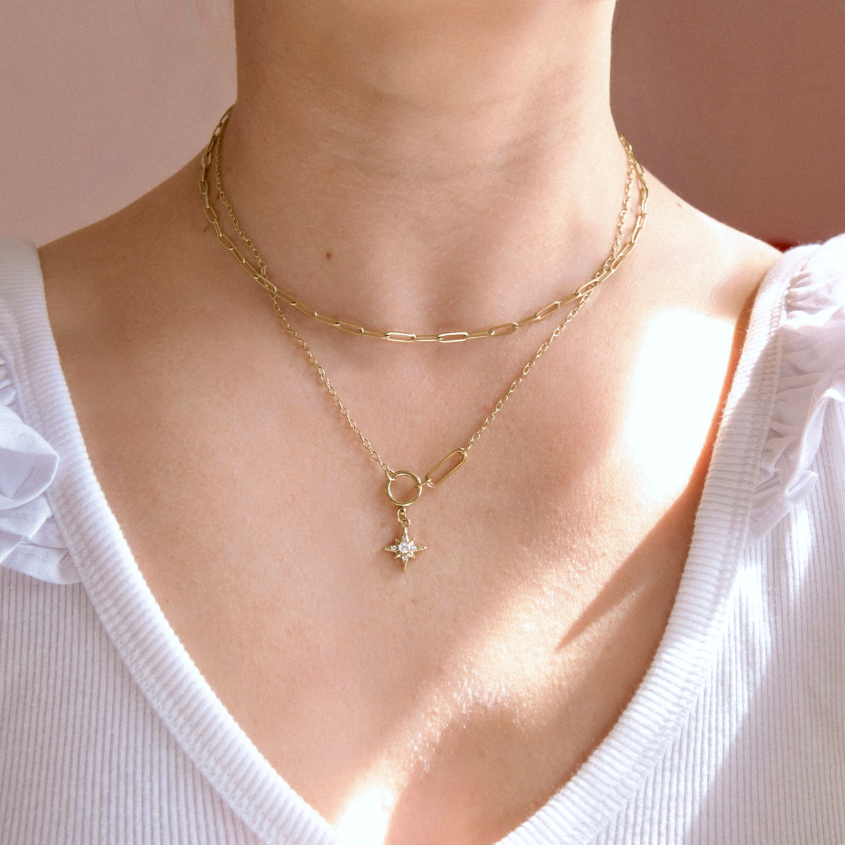 A close-up of a person's neck and upper chest wearing layered gold necklaces, including a paperclip chain, a delicate chain with a small ring detail, and a star-shaped pendant, paired with a white top.