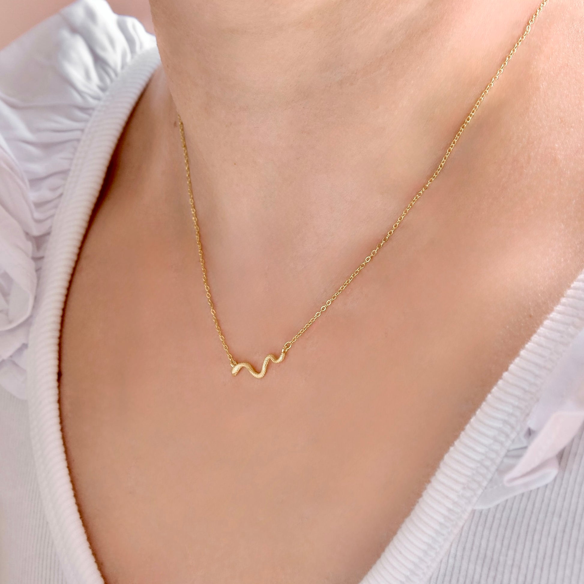 A close-up of a person wearing a delicate gold chain necklace with a small wavy pendant, styled with a white ribbed top.