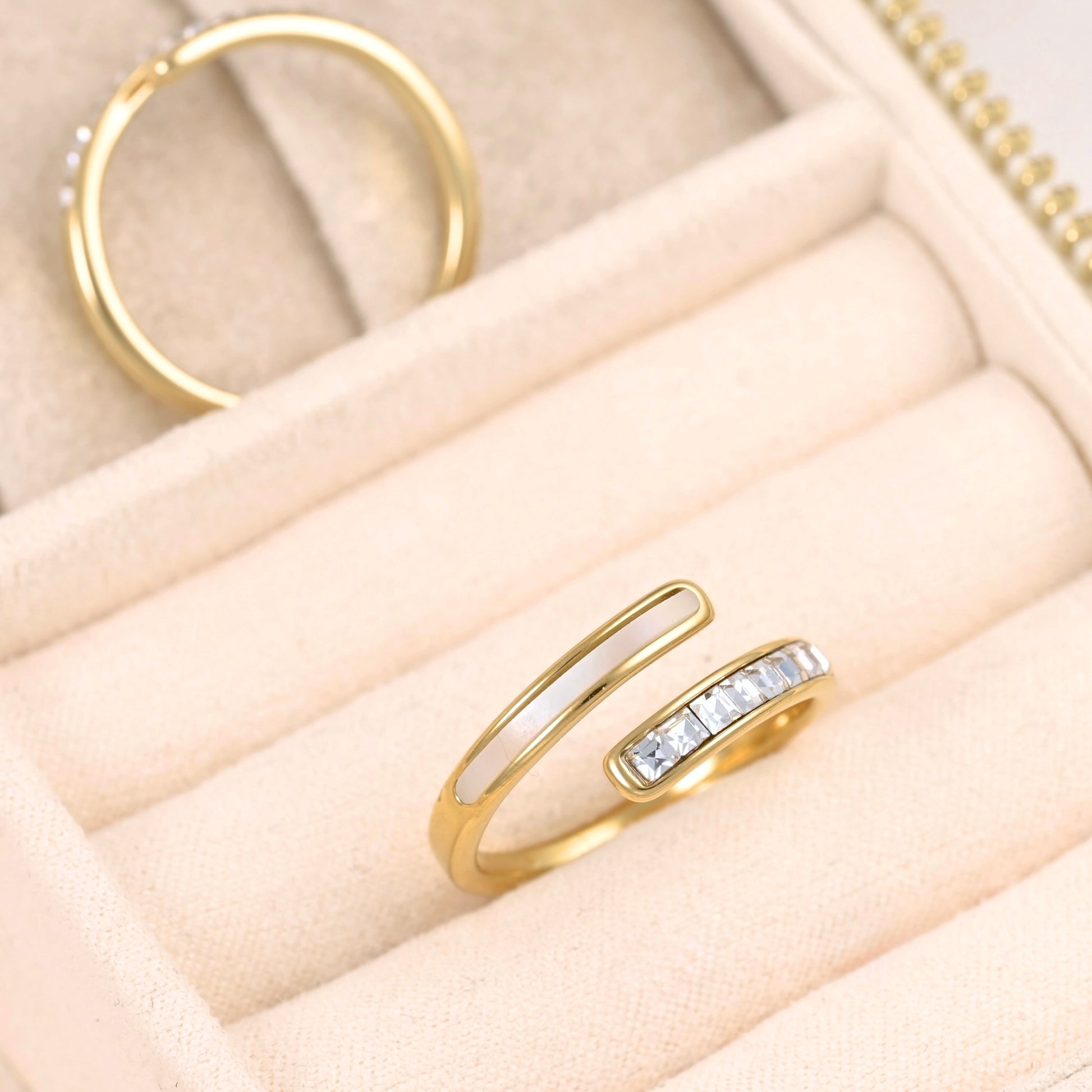 A gold open-band ring with a white inlay and a row of clear stones on one side, displayed in a beige jewelry box. Another gold ring is partially visible in the background.