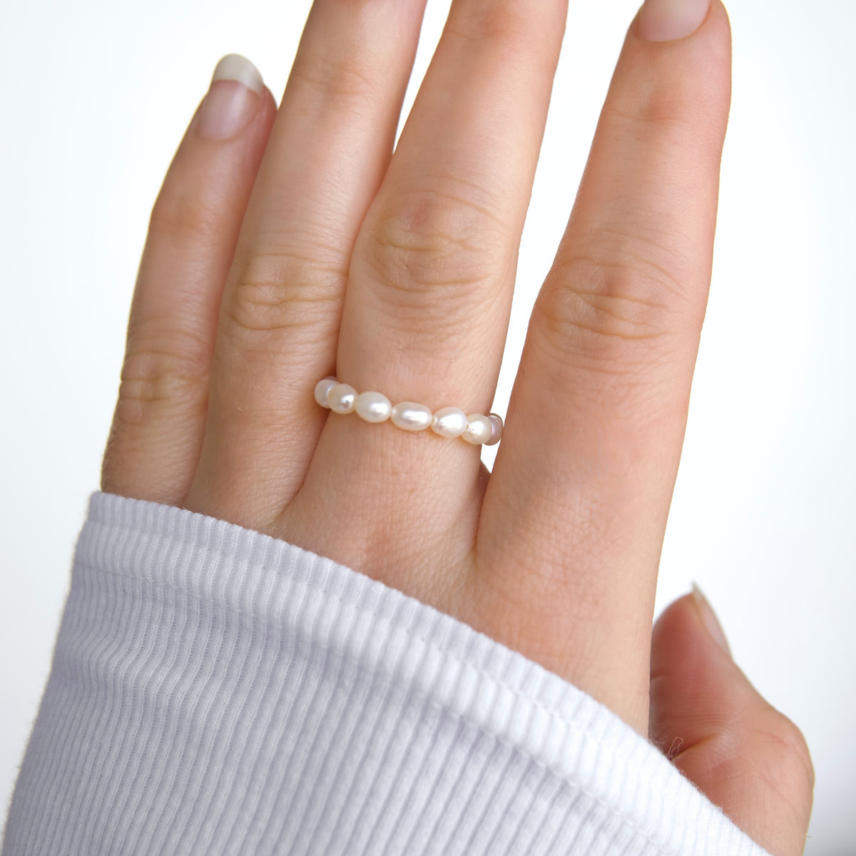A close-up of a hand wearing a delicate ring made of small white pearls, with a white ribbed long-sleeve shirt visible against a plain white background.