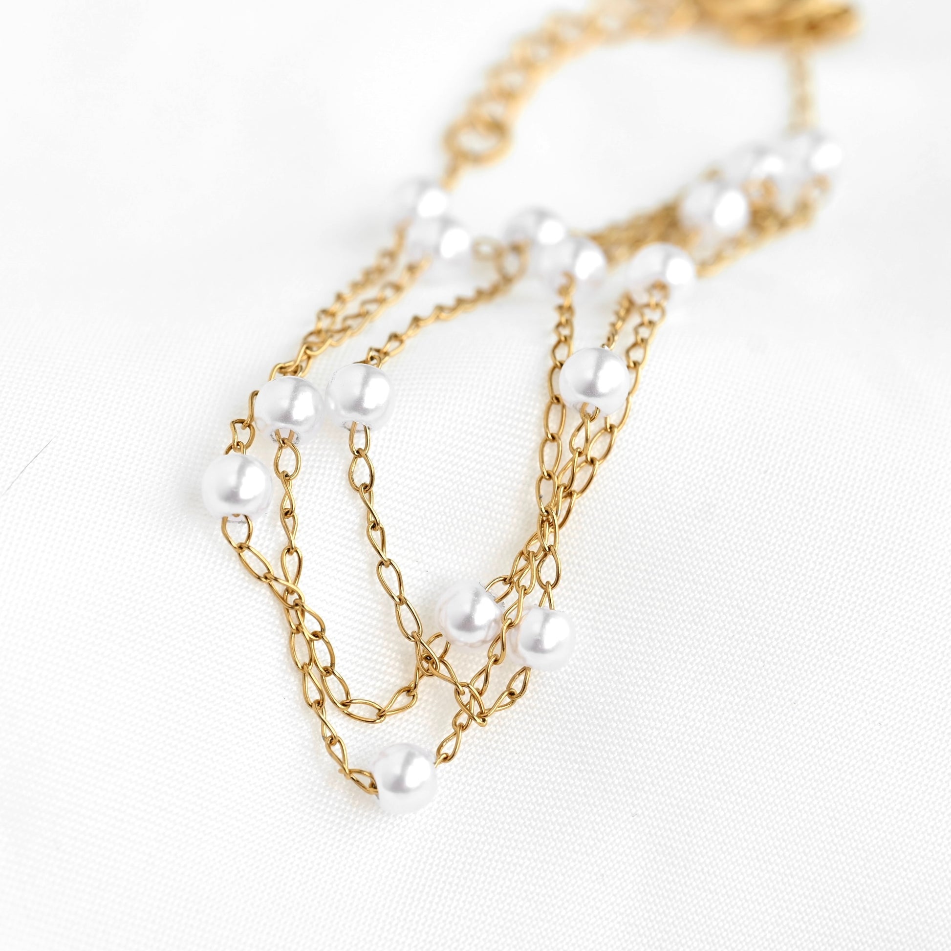 A gold chain necklace adorned with small white pearls, displayed on a textured white fabric background.
