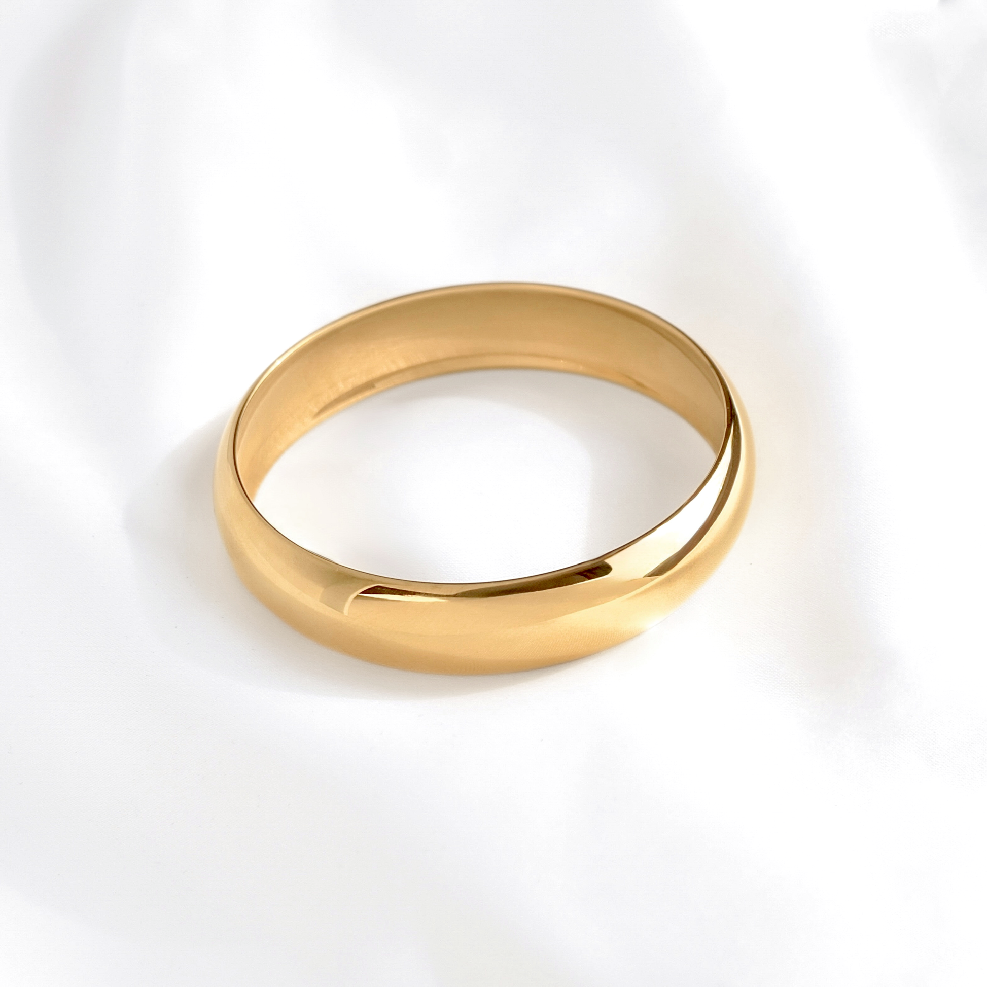 A polished yellow gold bangle with a unique curved, open design, resting on a soft white fabric background.