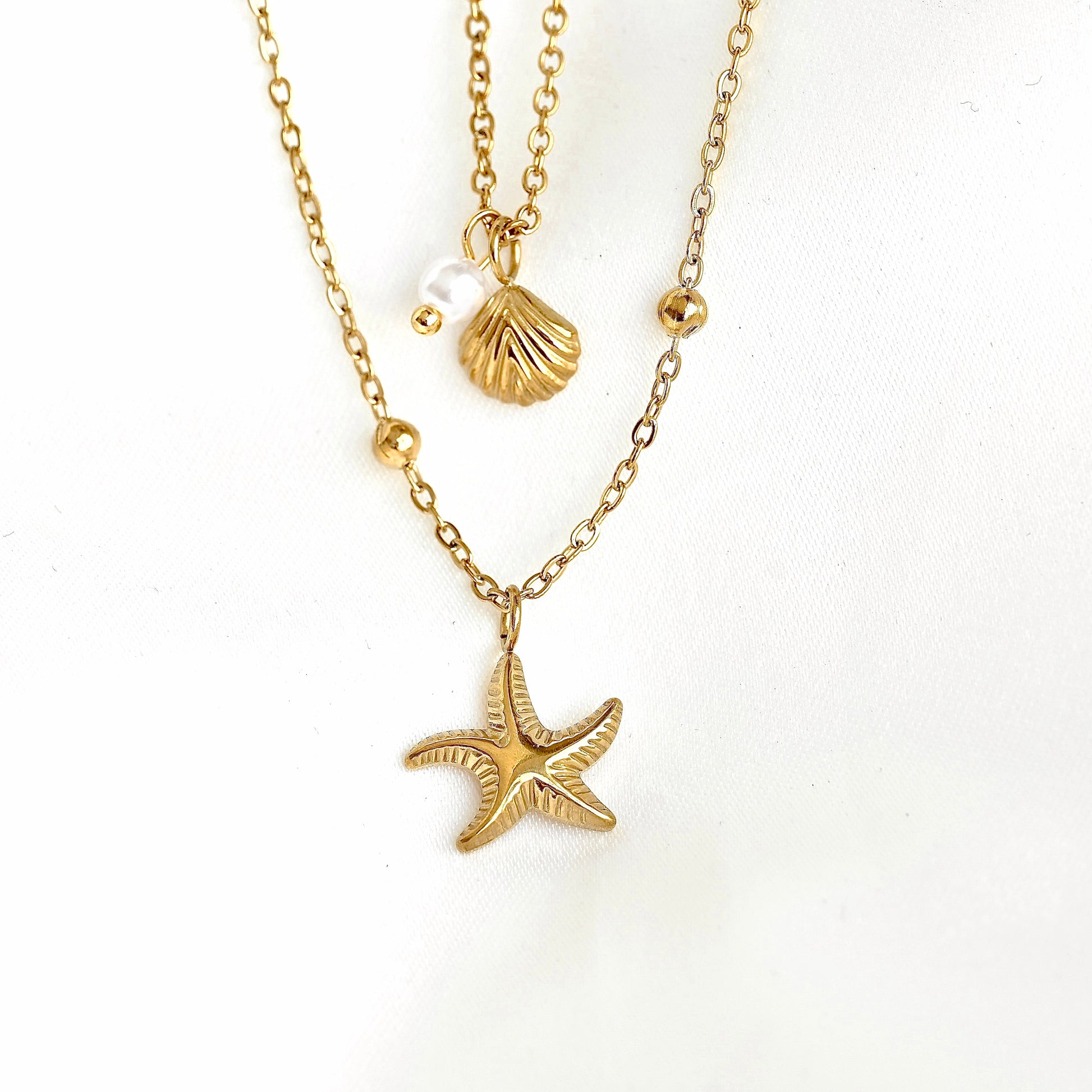 A gold-tone layered necklace featuring a starfish pendant, a seashell charm, and small gold beads on a white background.