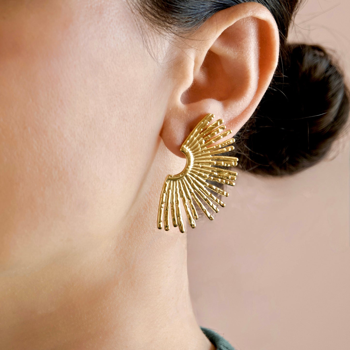 A close-up of a model's ear wearing a gold crescent moon earring with radiating sunburst prongs, set against a soft beige background.