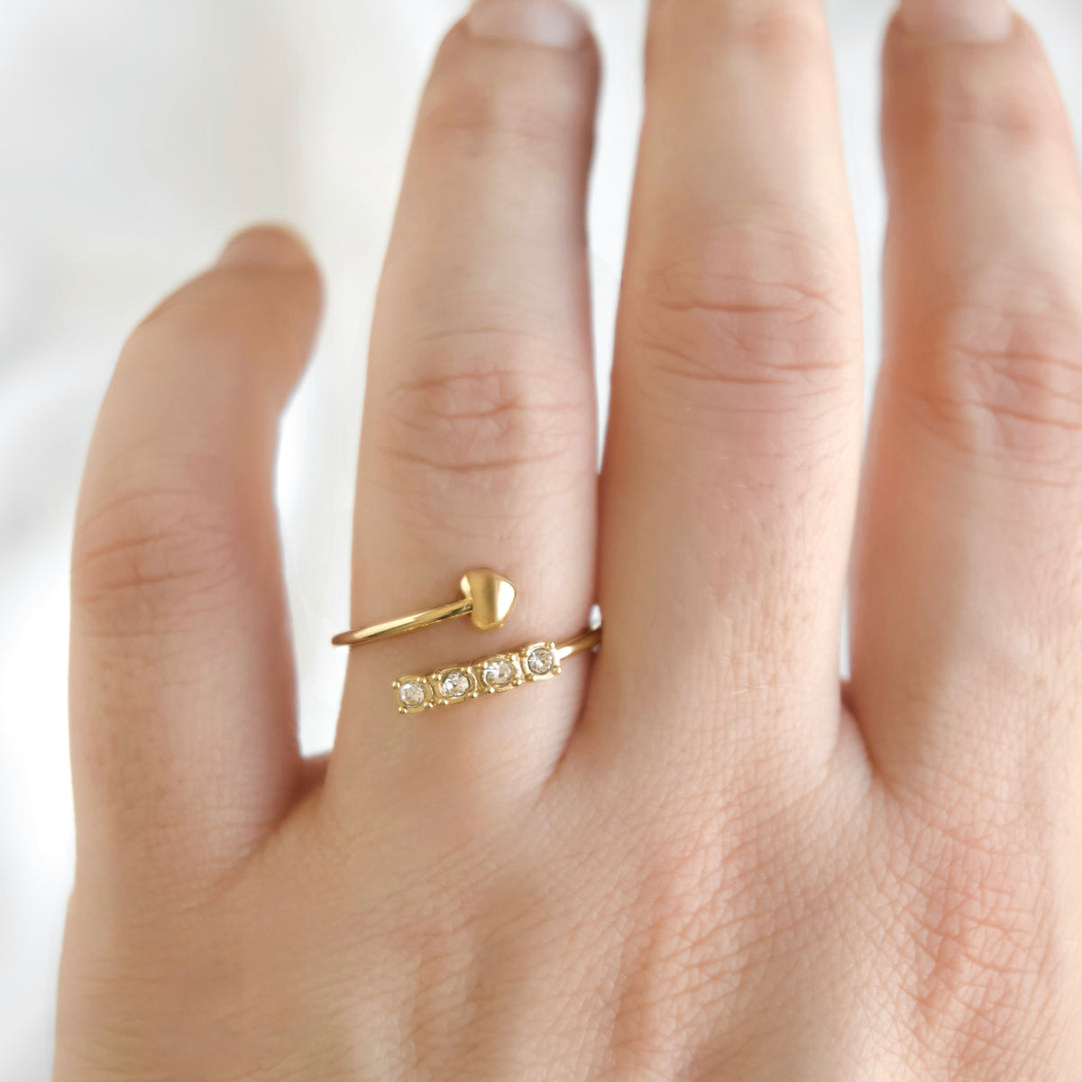 A close-up of a hand wearing a delicate gold ring featuring a small heart-shaped top and a row of small clear stones, set against a soft white background.