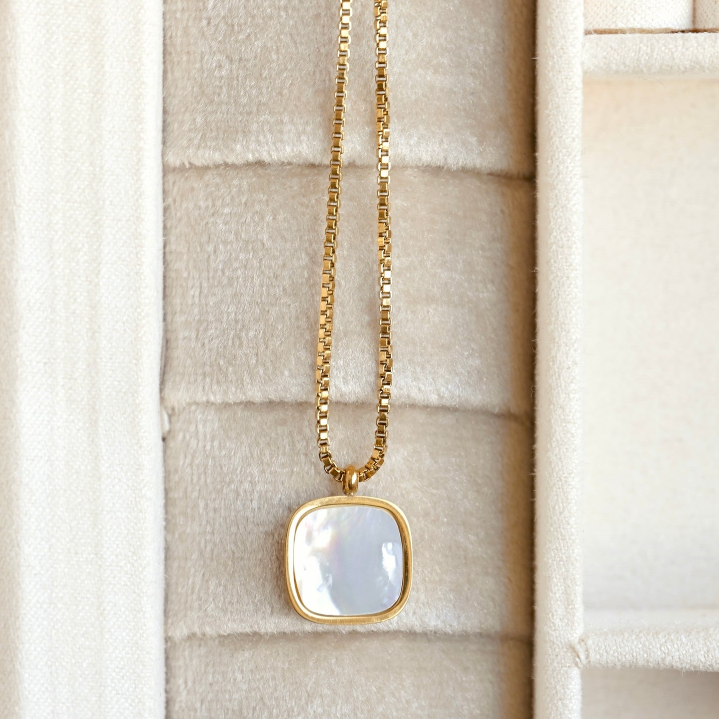 A gold chain necklace with a square bezel pendant featuring a white mother-of-pearl inlay, displayed inside a cream-colored jewelry box.
