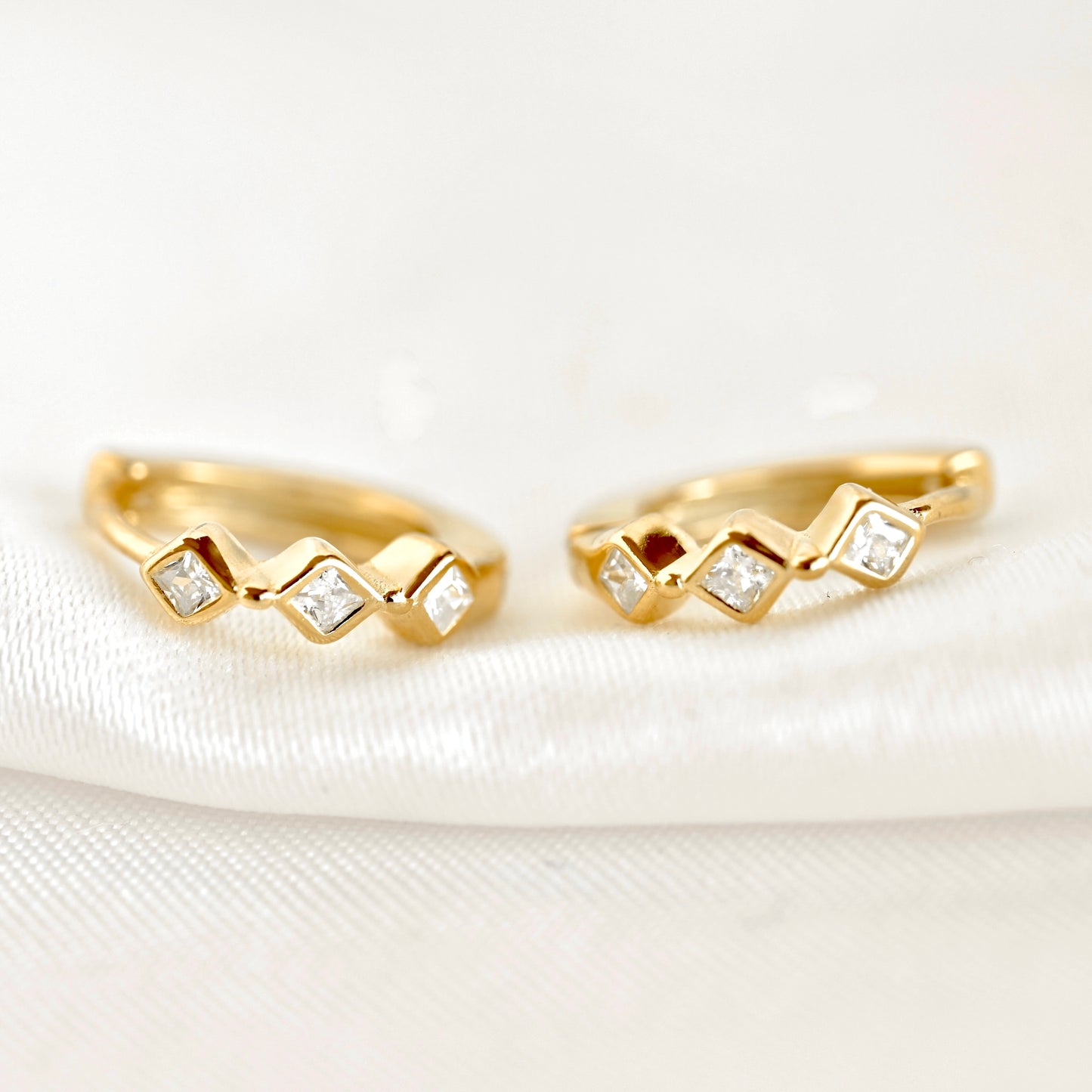 A pair of gold huggie hoop earrings with a zigzag design, each set with small clear square-cut gemstones, resting on a white textured fabric background.
