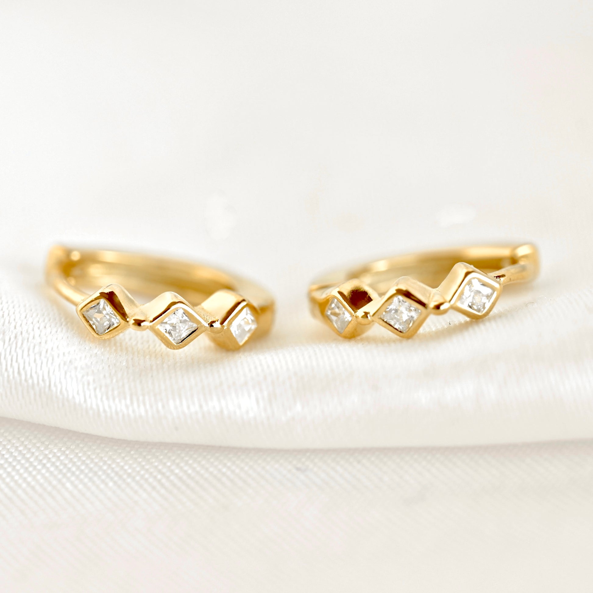 A pair of gold huggie hoop earrings with a zigzag design, each set with small clear square-cut gemstones, resting on a white textured fabric background.
