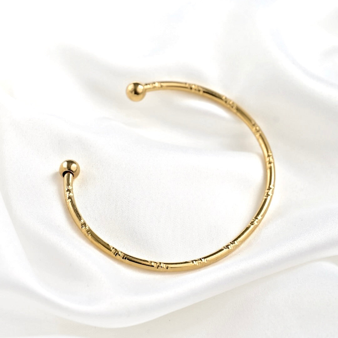 Top view of a gold-toned open cuff bracelet with a textured, segmented look and polished ball ends, displayed on a soft white satin fabric background.