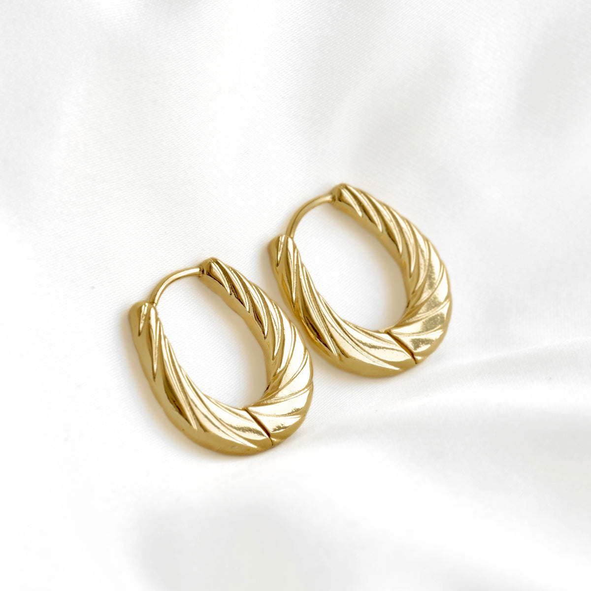 A pair of gold hoop earrings with a twisted, ribbed texture, displayed on a soft white fabric background.