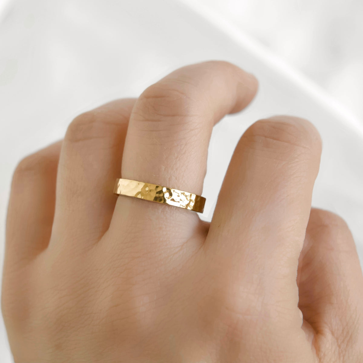 A close-up of a hand wearing a thin hammered gold band ring, set against a soft, bright white background.