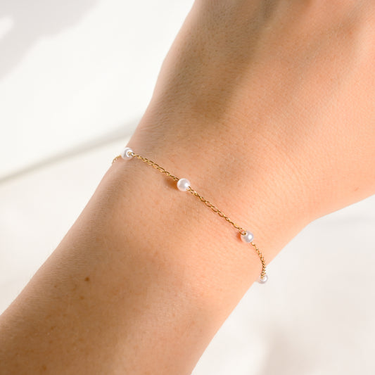 A delicate gold chain bracelet with three small white pearls, shown worn on a person's wrist against a bright, soft-focus background.