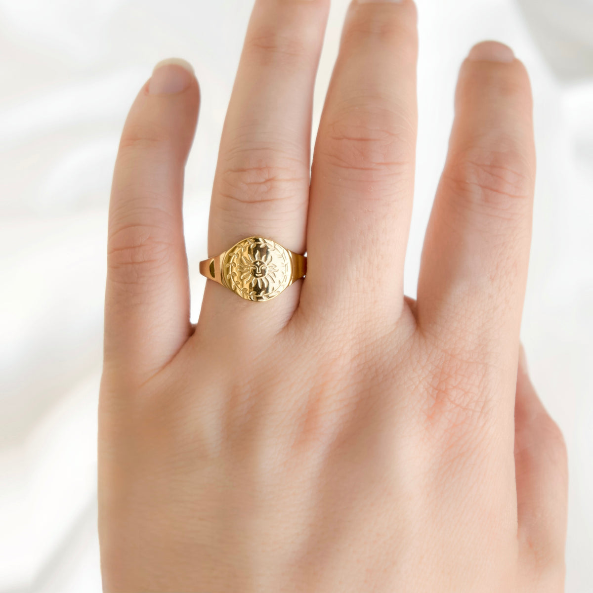 A close-up of a hand wearing a textured gold signet ring with a circular sun design on a soft white background.