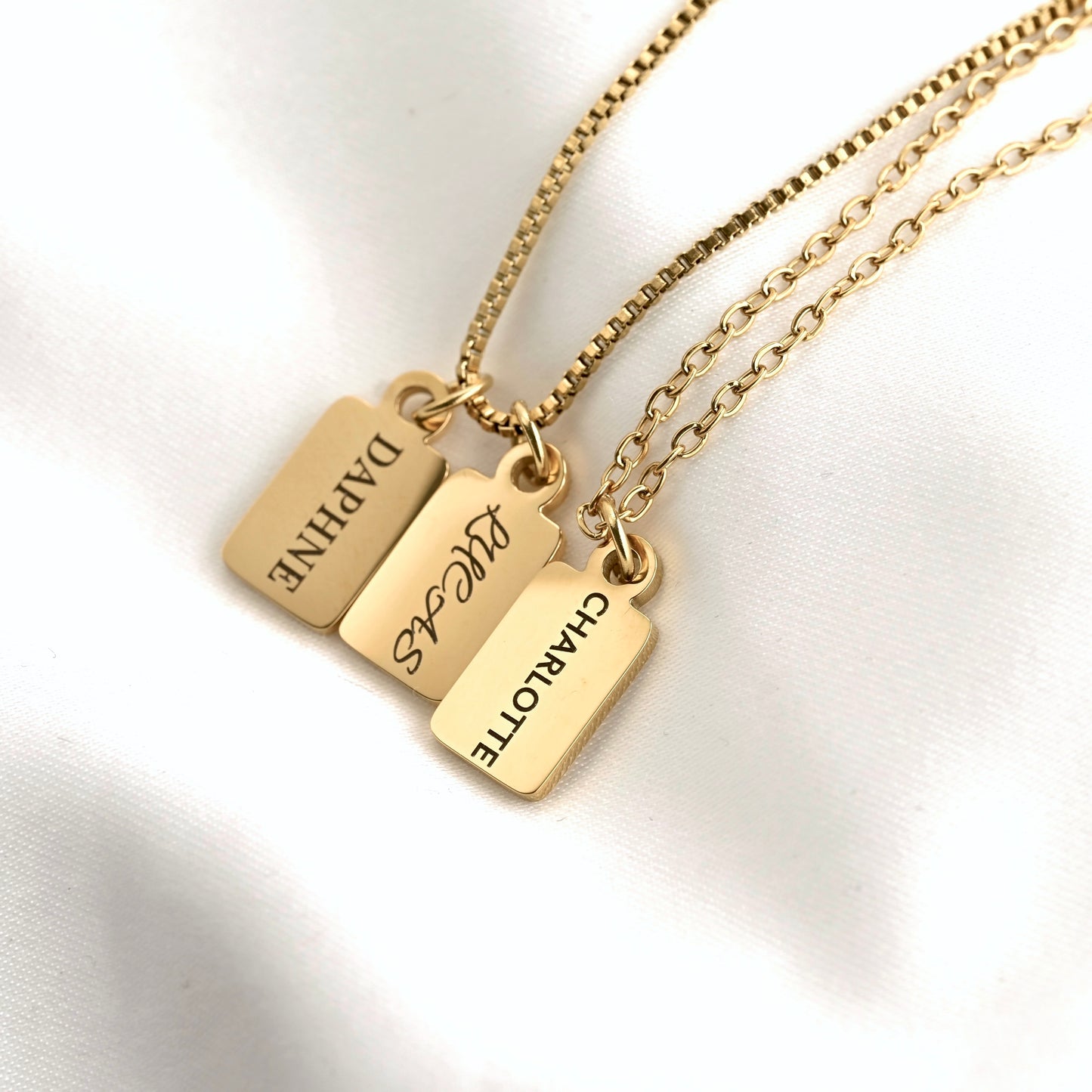 Three gold nameplates on chains, personalized with the names DAPHNE, LUCAS, and CHARLOTTE, displayed on a white fabric background.