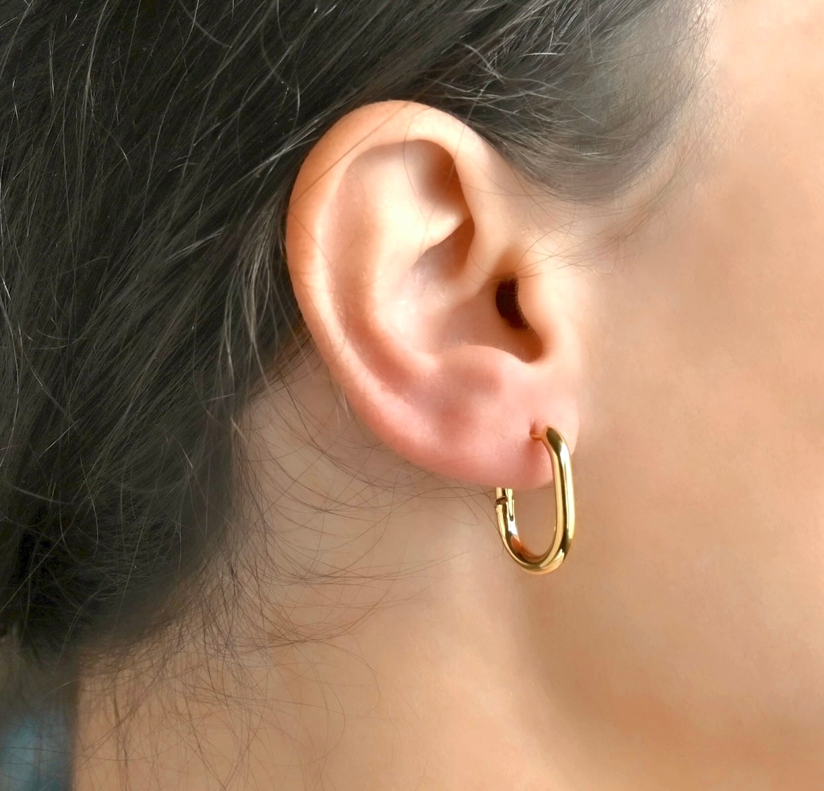 Hinged elongated oval hoop earrings in gold on a person's ear