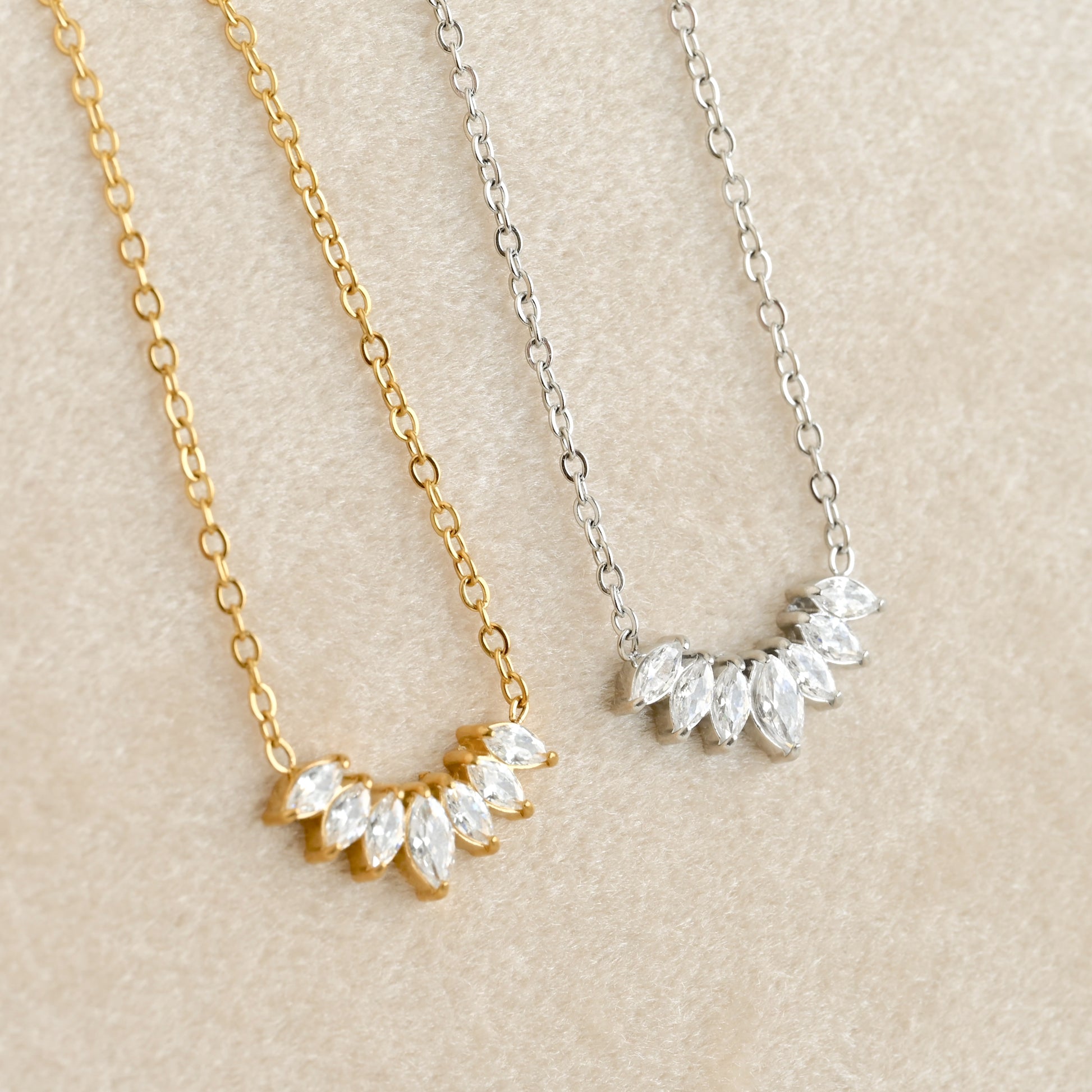 Two delicate necklace pendants on a beige textured background, one in yellow gold and one in silver, each featuring a curved row of clear marquise-cut stones.