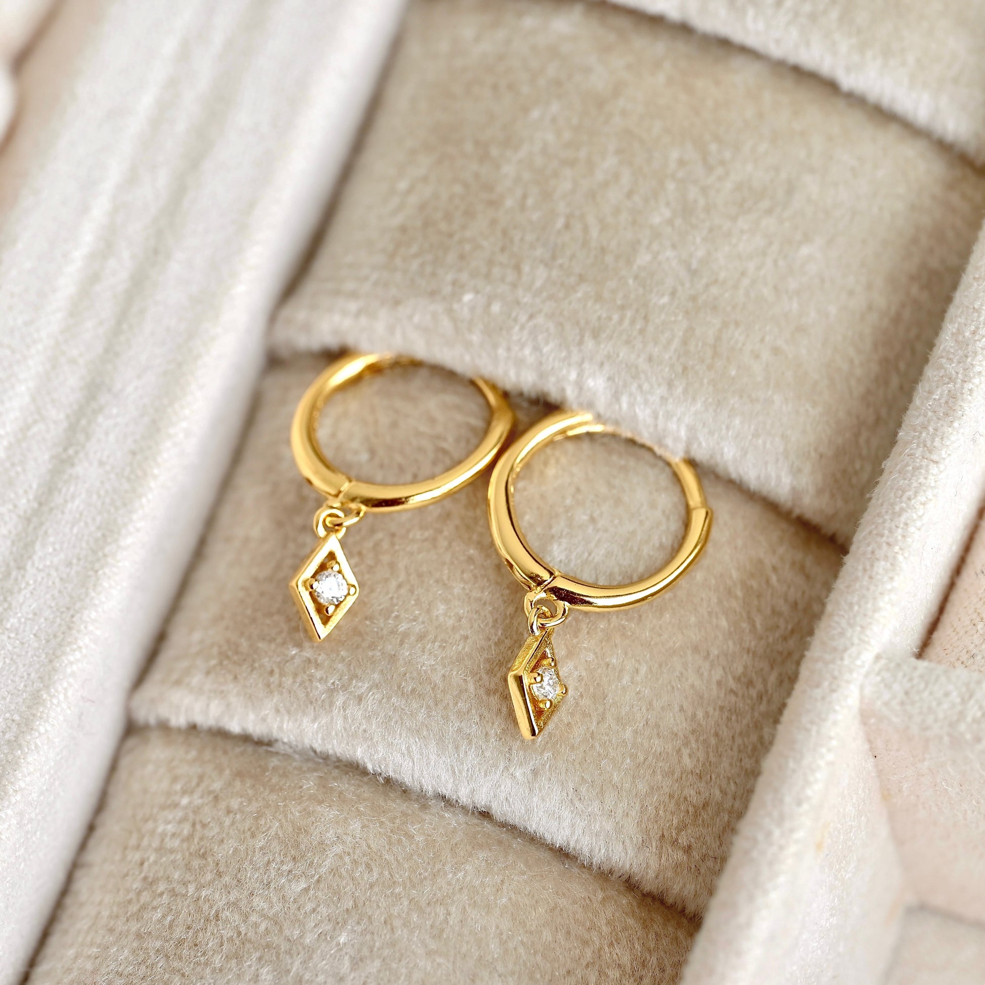 A pair of gold huggie hoop earrings with dangling geometric diamond-shaped charms set with small clear stones, displayed on a beige velvet jewelry box.