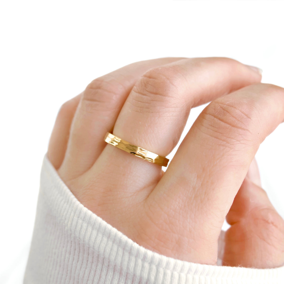 A close-up of a hand wearing a thin, etched gold band ring, with the hand resting against a white ribbed sleeve and a clean white background.