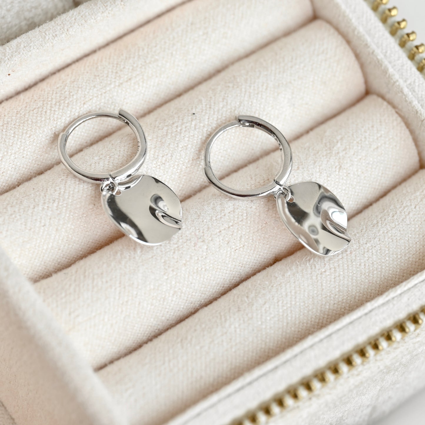A pair of silver huggie hoop earrings with polished, organic-shaped dangling charms, displayed on a cream-colored jewelry box cushion.