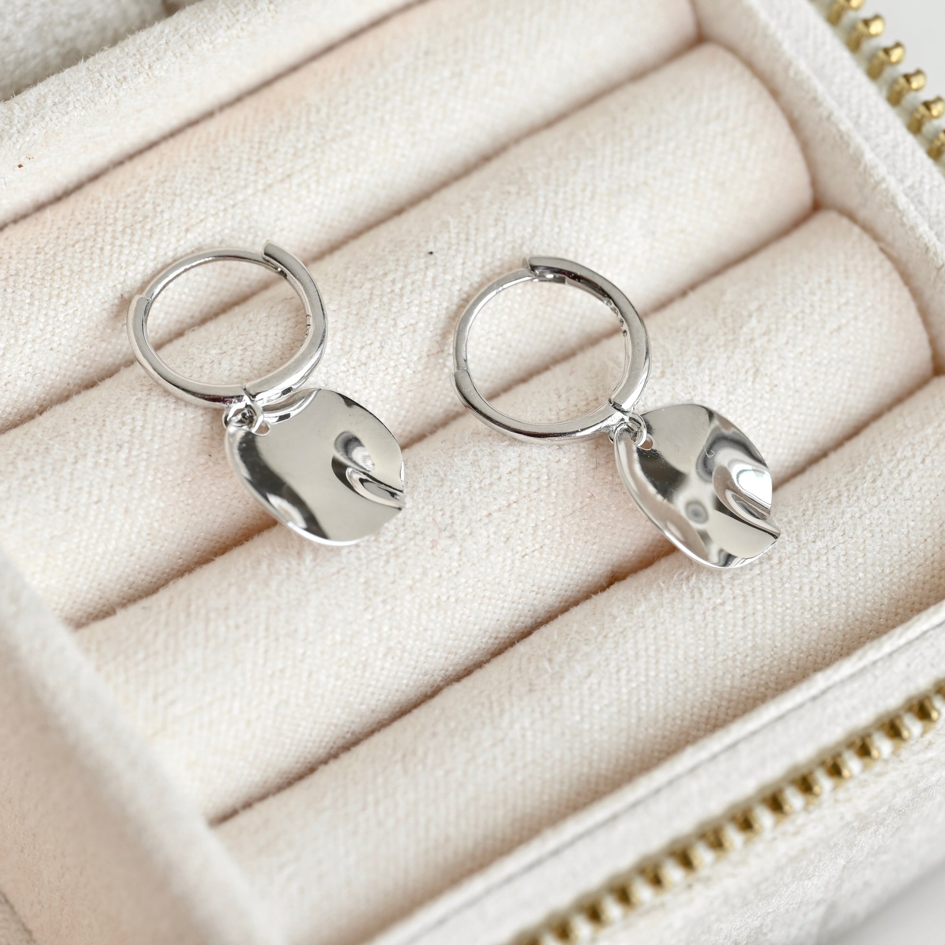 A pair of silver huggie hoop earrings with polished, organic-shaped dangling charms, displayed on a cream-colored jewelry box cushion.