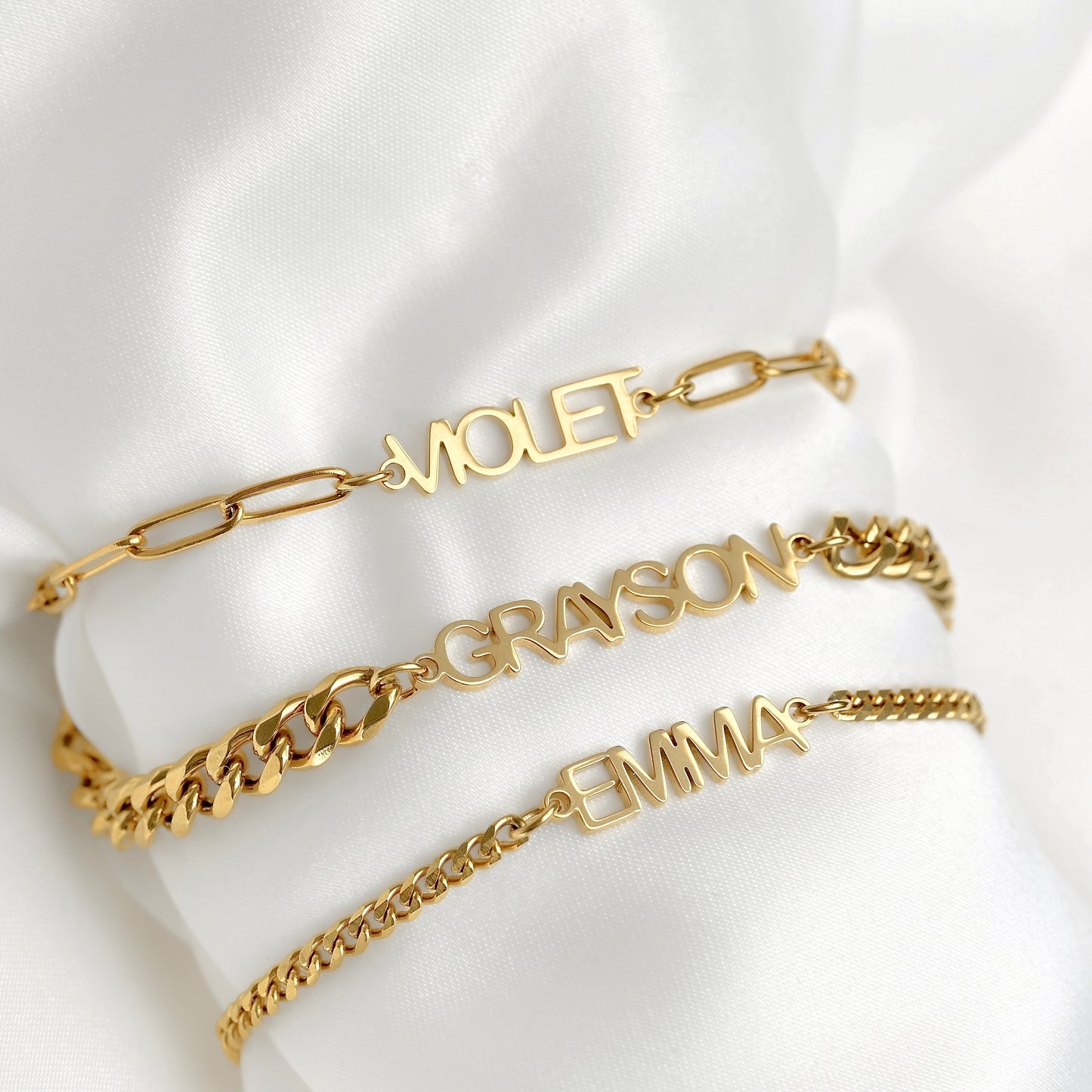Three gold name bracelets with different chain links are displayed on a white satin fabric. The top bracelet has paperclip links and the name 'MIOLET', the middle has curb links and 'GRAYSON', and the bottom has curb links and 'EMMA'.