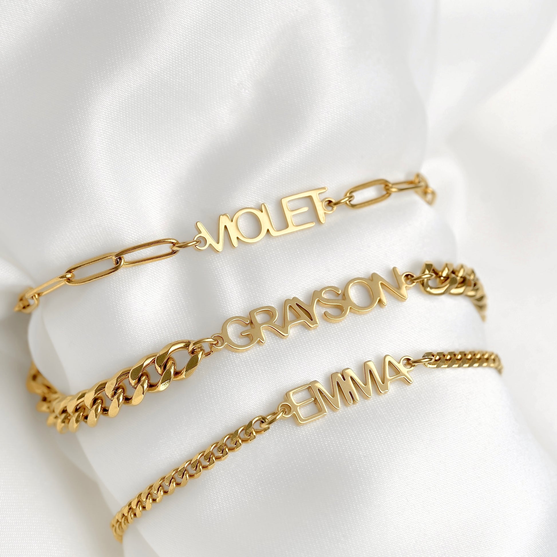 Three gold name bracelets with different chain links are displayed on a white satin fabric. The top bracelet has paperclip links and the name 'MIOLET', the middle has curb links and 'GRAYSON', and the bottom has curb links and 'EMMA'.