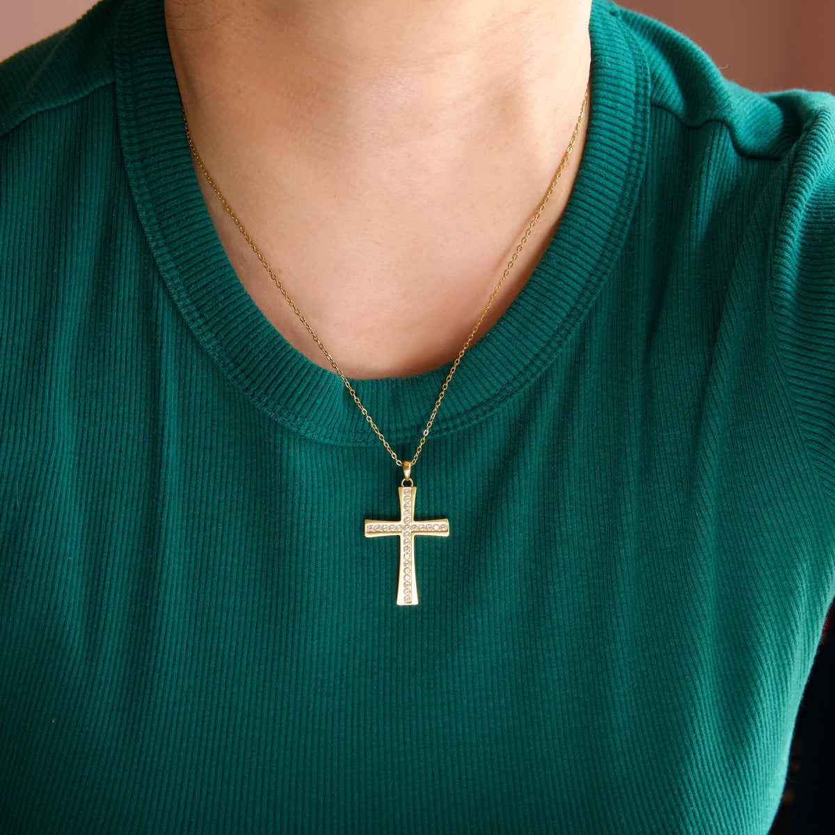 A gold cross pendant necklace with clear stones, worn by a person wearing a dark green ribbed t-shirt.