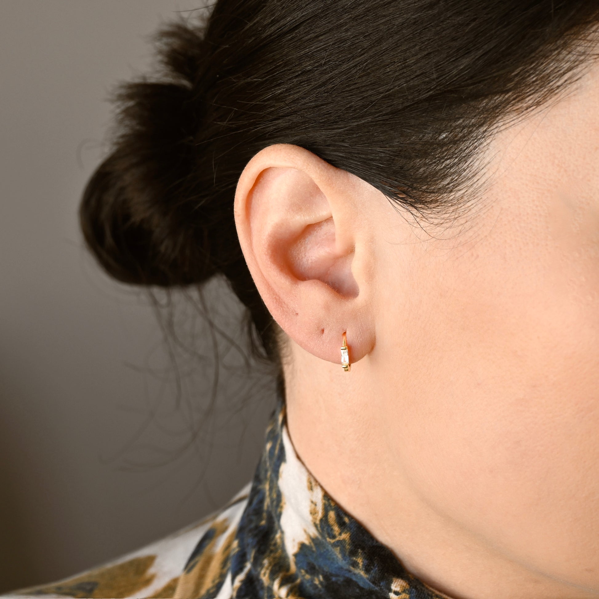A close-up of a person's ear wearing small gold hoop earrings with a clear rectangular cubic zirconia, against a neutral gray background.