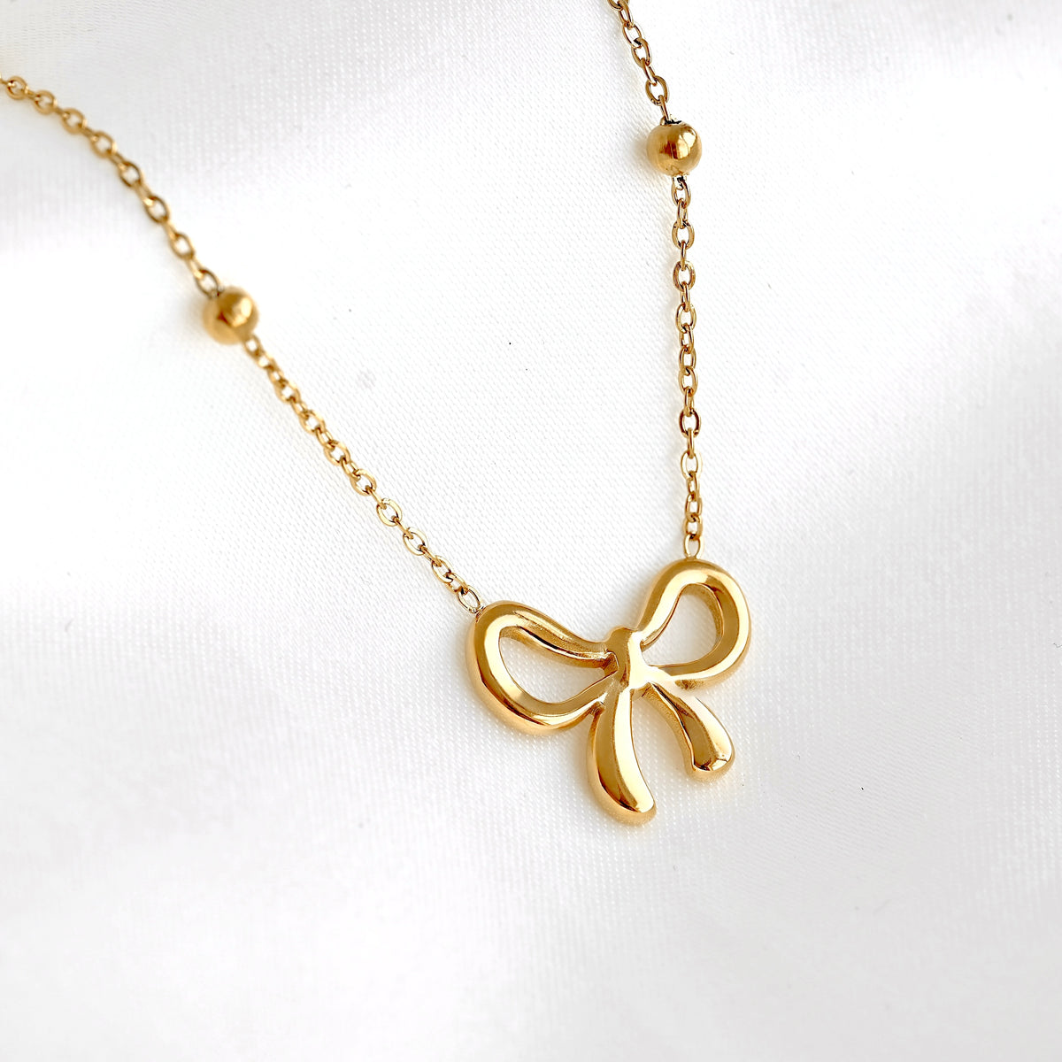 A delicate gold chain necklace featuring a polished bow-shaped pendant and two small gold beads spaced along the chain, displayed on a soft white fabric background.