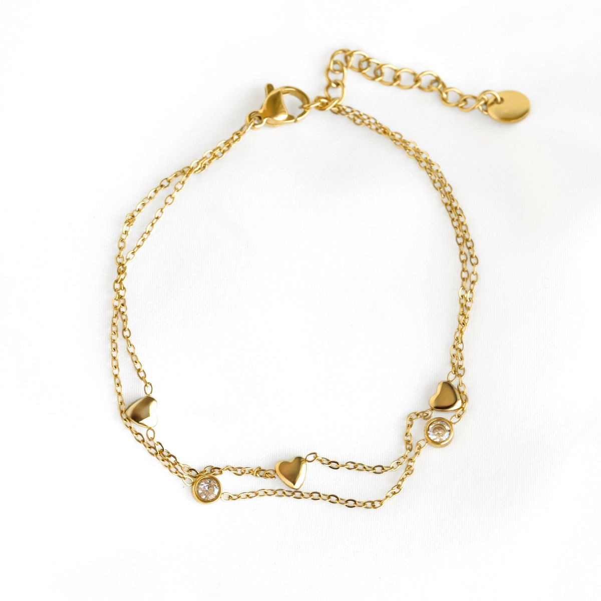 A delicate gold double-chain bracelet featuring small heart charms, one of which is set with a clear crystal, displayed on a clean white background.