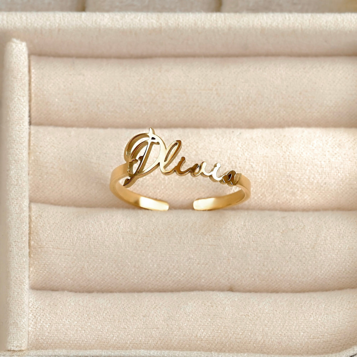 A gold script name ring spelling 'Olivia' resting on a beige velvet jewelry display tray.