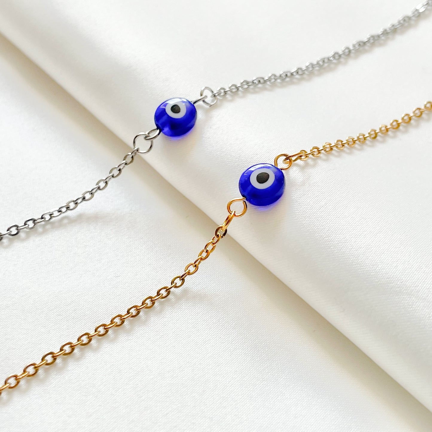 Two bracelets, one gold one silver, with blue evil eye beads on a white background