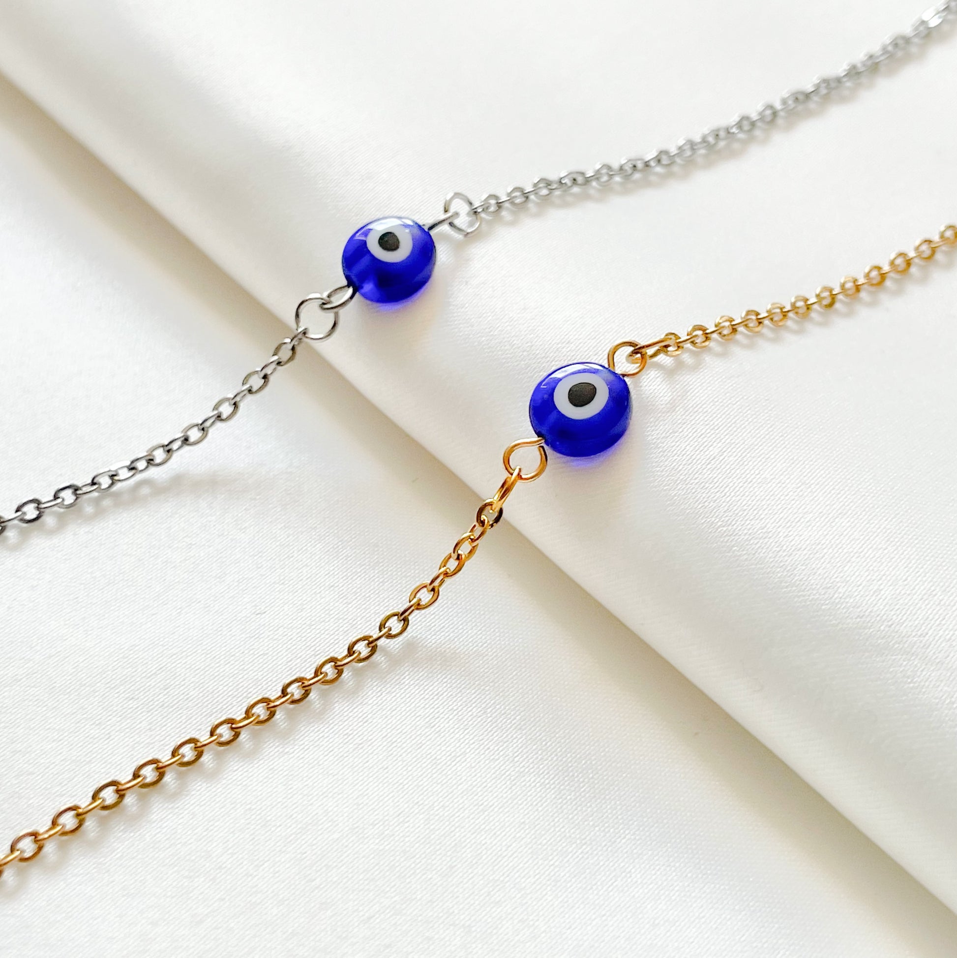 Two bracelets, one gold one silver, with blue evil eye beads on a white background