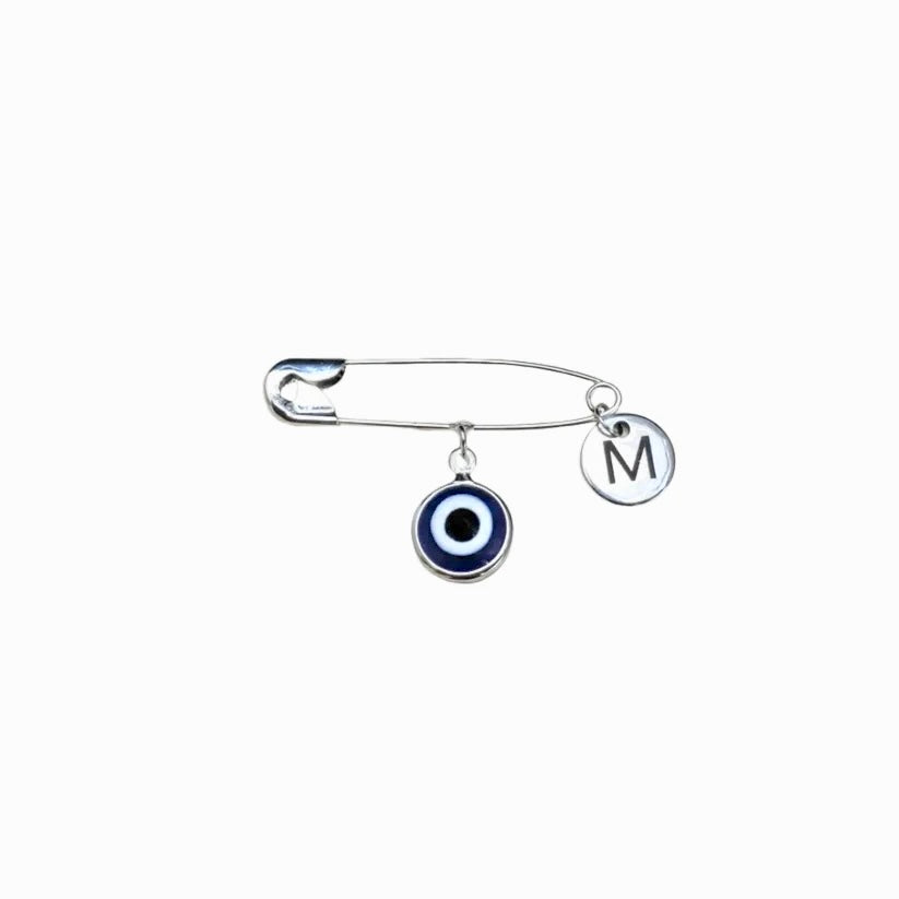 Silver safety pin with a blue evil eye charm and 'M' charm on a white background