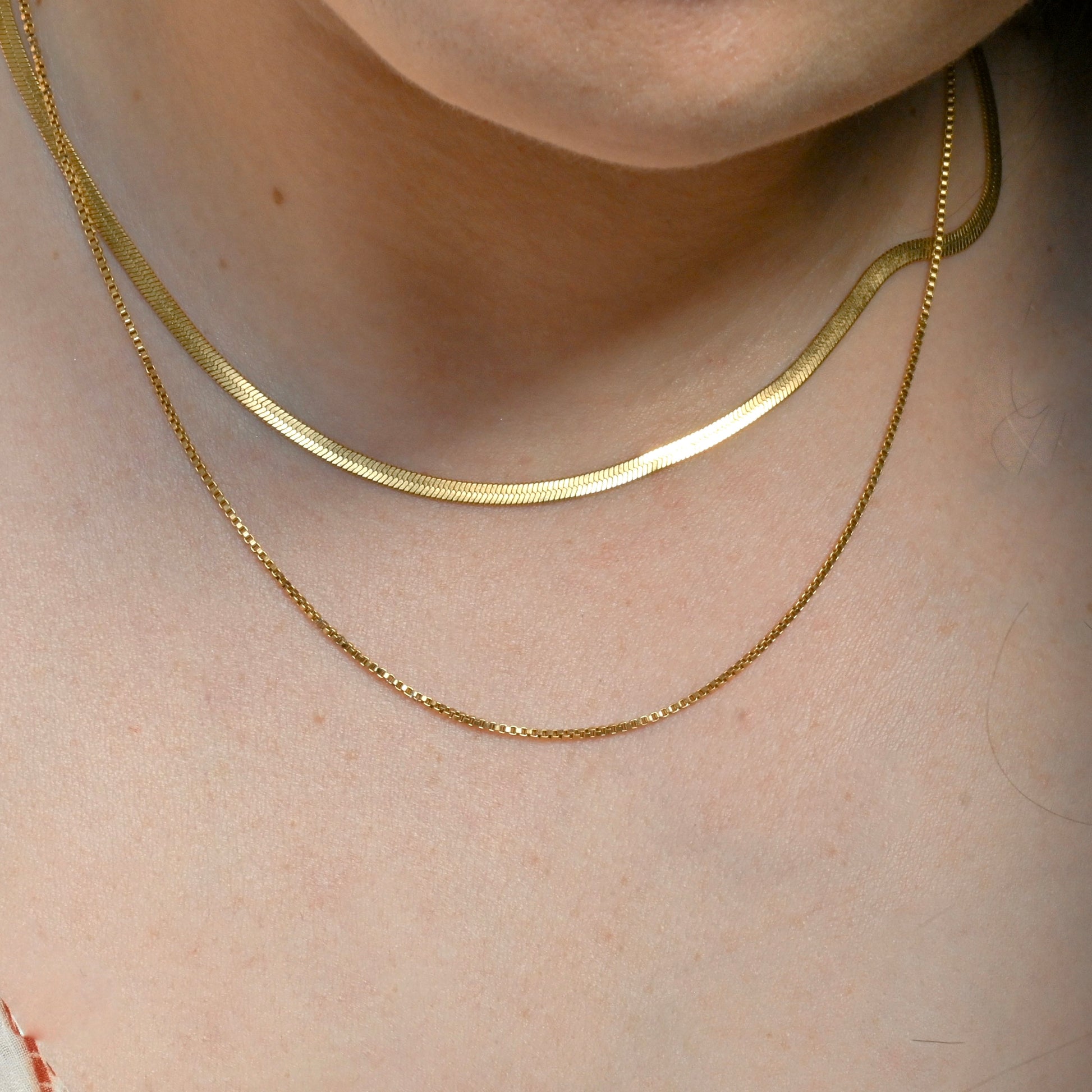 Gold necklace set with herringbone and box chain on a person's neck with a neutral background