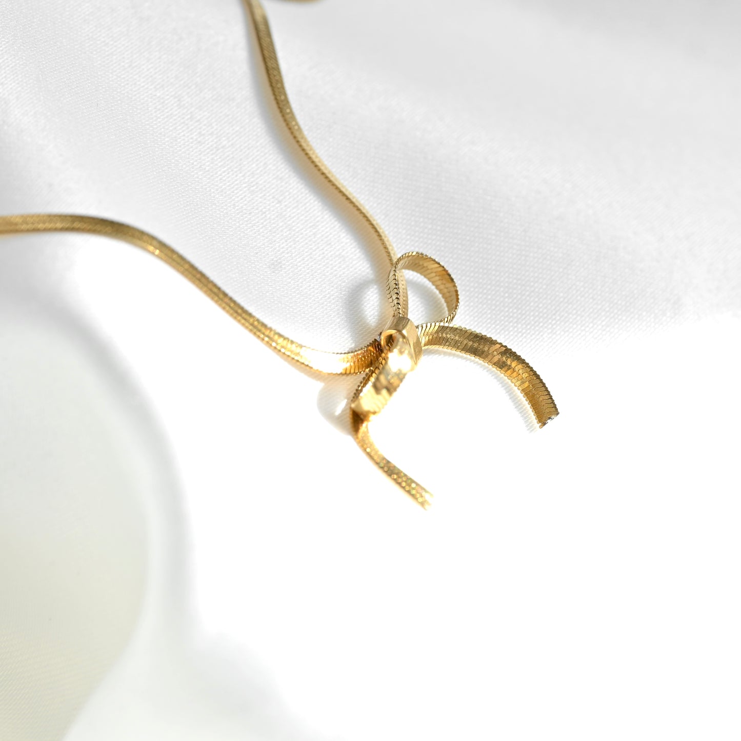 A gold chain necklace with a textured bow pendant, displayed on a white satin fabric background.