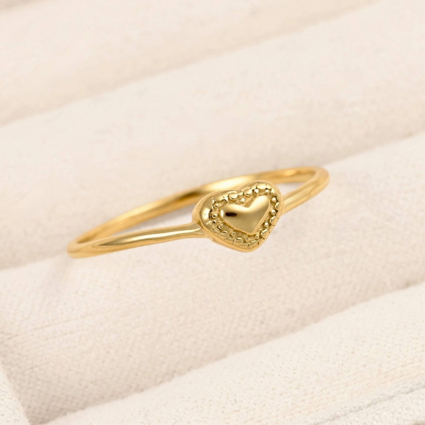 A delicate gold ring featuring a small heart-shaped charm with a beaded border, resting on a textured cream-colored ring box.
