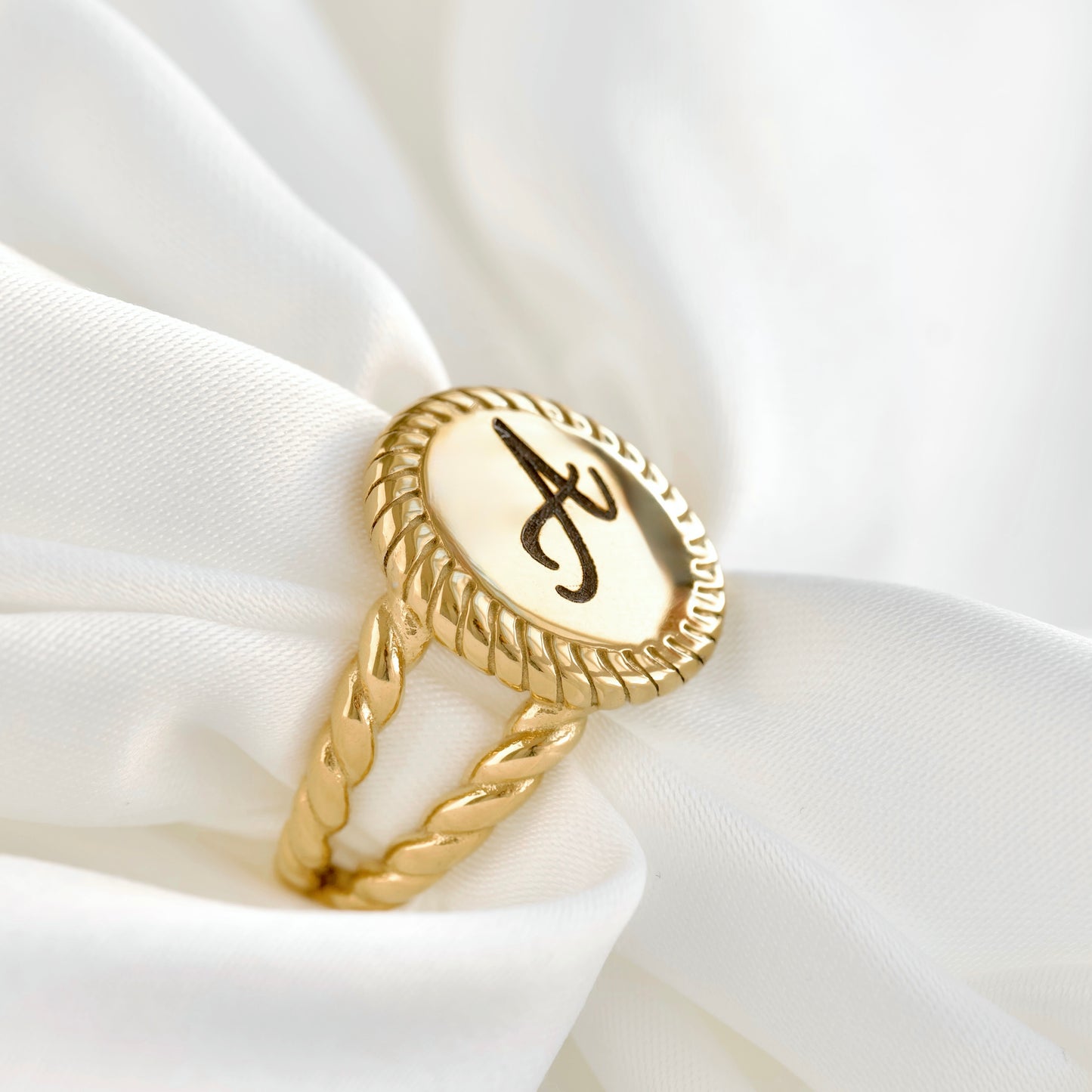 A gold ring with a twisted rope-style band and an oval face featuring a black monogram letter 'A' on a cream background, displayed on draped white fabric.